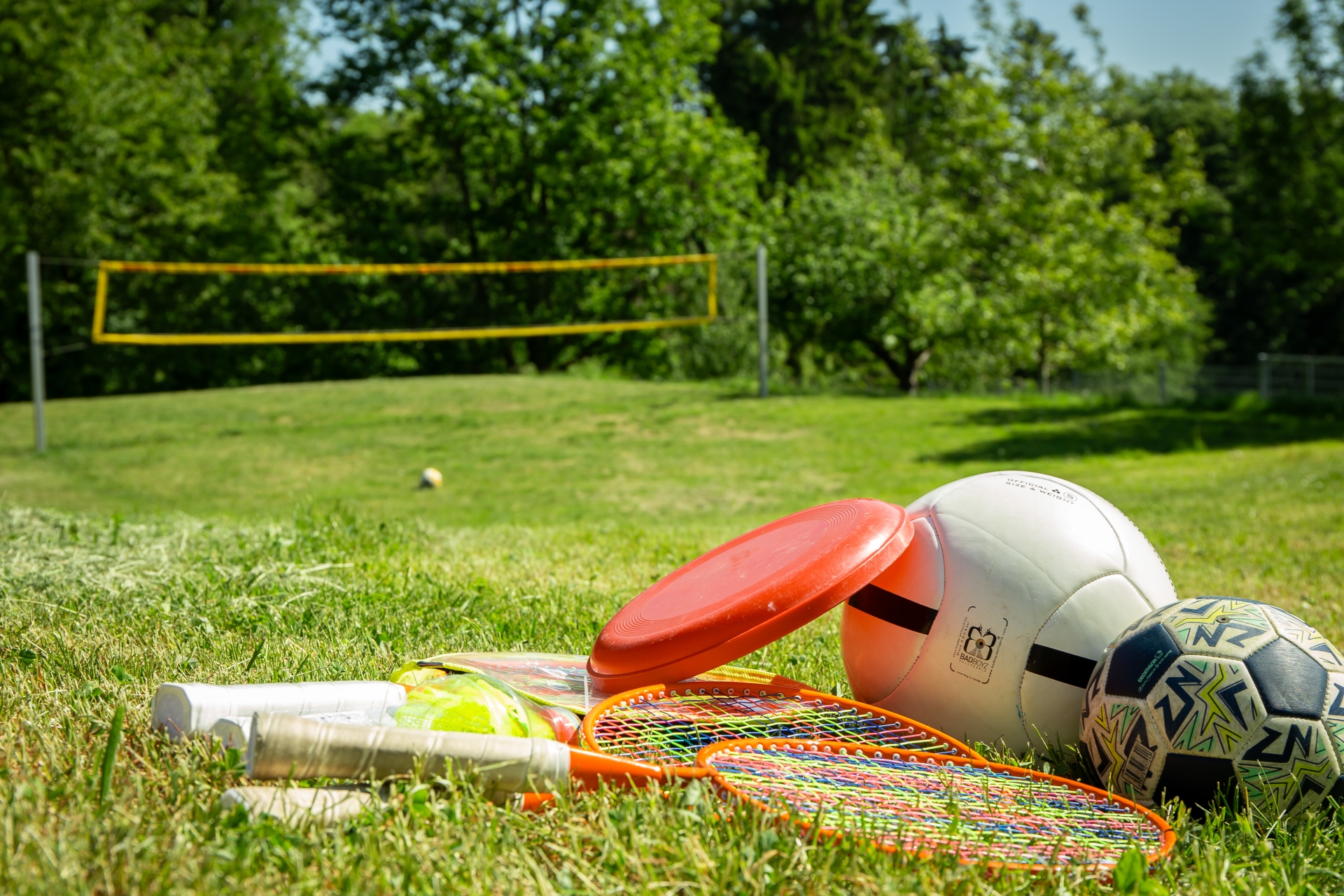 Mehrere Sportgeräte liegen auf einer Wiese: zwei Badmintonschläger mit Federbällen, eine orangefarbene Frisbee-Scheibe, ein Volleyball und ein Fußball. Im Hintergrund ein Volleyballnetz und Bäume.