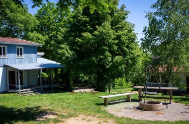 Blaues Holzhaus mit Veranda neben mehreren Bänken und einem runden Feuerplatz mit Dreibeingrill, umgeben von grünen Bäumen.