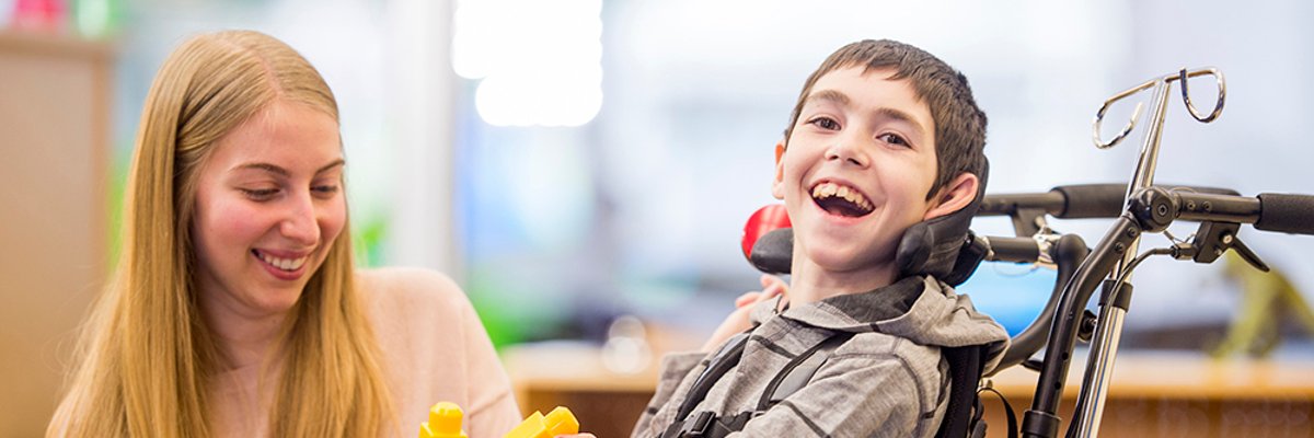 Foto: Lachender Junge im Rollstuhl mit junger Frau als Assistenzkraft.