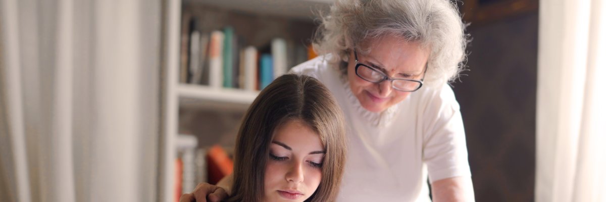 Foto: Ältere Frau steht hinter einem Kind und schaut ihm über die Schulter, während es in einem Buch liest.