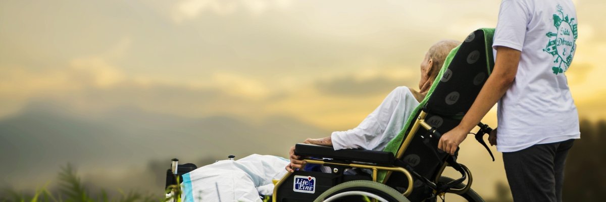 Foto: Eine Pflegerin und ein Mann in einem Liegerollstuhl stehen auf einer Wiese und betrachten den Sonnenuntergang.