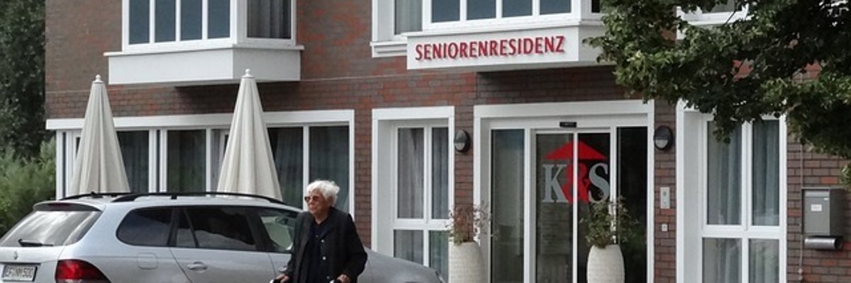 Foto: Eingang eines Seniorenheims. Davor ein alter Mann mit Rollator.