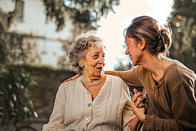 Foto: Eine ältere und eine jünger Frau sitzen nebeneinander. Die jüngere hat den Arm um die Schultern der älteren gelegt. Mit der anderen Hand hält sie die Hand der älteren. Die beiden lachen sich an.