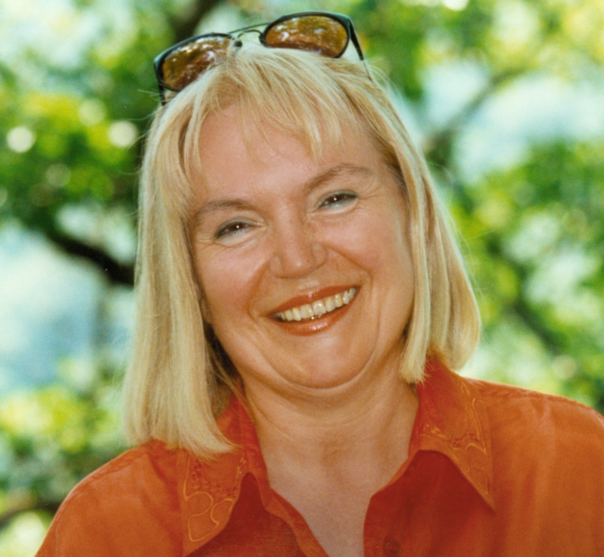 Das Foto zeigt Dr. Dagmar Morgan. Sie lacht, hat eine positive Ausstrahlung. trägt eine orange Bluse. Im Hintergrund ist eine Baumkrone.