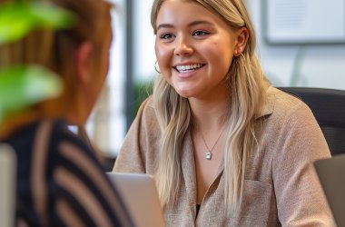 Zwei Frauen sitzen sich am Schreibtisch gegenüber. Die Frau im Vordergrund lächelt freundlich und trägt eine beige Bluse, während sie auf das Gegenüber eingeht. (KI-generiert)