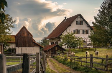 Ländlicher Hof mit Fachwerkhaus, Scheune und eingezäunter Weide, auf der mehrere Tiere grasen; ein Weg führt durch die grüne Landschaft zum Haus.(KI-generiert)