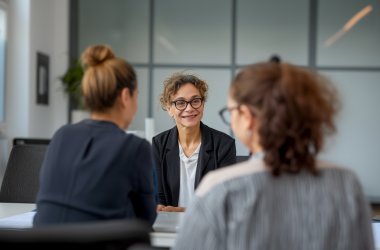 Drei Personen sitzen in einem Büro zusammen; eine Frau mit Brille spricht lächelnd mit zwei gegenüber sitzenden Kolleginnen.(KI-generiert)