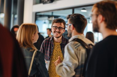 Ein Mann mit Brille lächelt einem anderen Mann zu, der mit dem Rücken zur Kamera steht. Eine Frau steht ebenfalls mit dem Rücken zur Kamera neben ihnen. Um sie herum stehen weitere Personen, die nicht an der Konversation beteiligt sind.(KI-generiert)