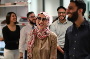 Fünf Personen im Arbeitskontext lachen gemeinsam. Im Vordergrund ist eine Frau zu sehen, die lächelt.(KI-generiert)