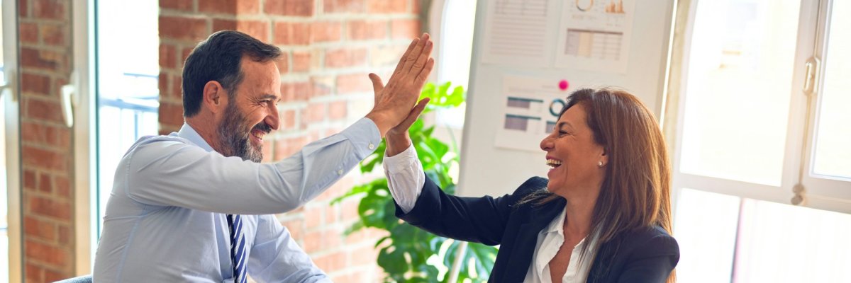 Auf dem Bild ist eine Frau und ein Mann im Büro zu sehen. Sie geben sich lachend einen Handschlag auf die gute Zusammenarbeit.