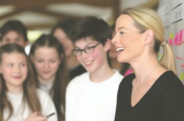 Referentin mit blondem Pferdeschwanz und schwarzem Oberteil steht vor einer Wand mit bunten Haftnotizen und einer Gruppe von Jugendlichen