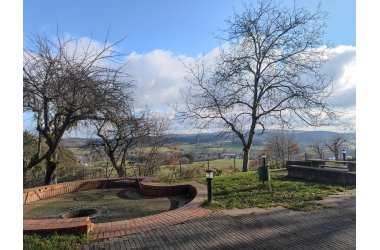 Blick auf eine gepflasterte Fläche auf der man grillen oder Lagerfeuer machen kann mit niedriger roter Mauer und einem grünen Mülleimer auf einer Wiese.