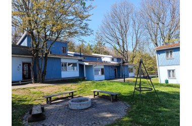 Mehrere blaue Wohngebäude mit weißen Fenstern umgeben eine gepflasterte Feuerstelle mit drei Holzbänken und einem Dreibein-Grillgestell auf einer Wiese.