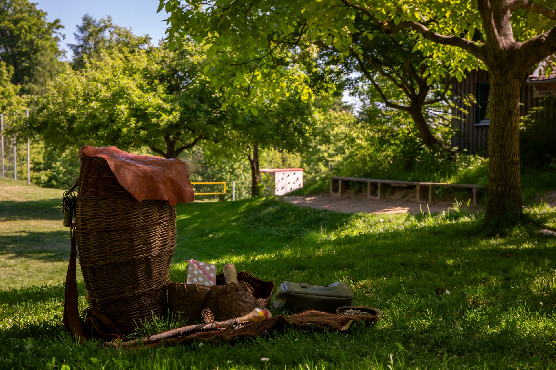 Großer, geflochtener Korb mit braunem Deckel steht auf Gras, daneben liegen mehrere kleine Körbe und Holzstücke, im Hintergrund Bäume und eine Bank.