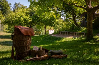 Großer, geflochtener Korb mit braunem Deckel steht auf Gras, daneben liegen mehrere kleine Körbe und Holzstücke, im Hintergrund Bäume und eine Bank.