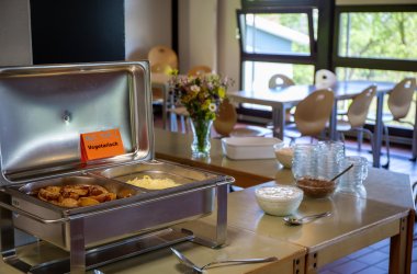 Offener Warmhaltebehälter mit vegetarischem Gericht, daneben Schalen mit verschiedenen Soßen auf einem Tisch in einem hellen Raum mit Stühlen und Fenster im Hintergrund.