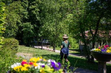 Frau mit Hut und dunkelblauer Jacke um die Taille geht auf einem Weg über eine grüne Wiese  mit bunten Blumen im Vordergrund.