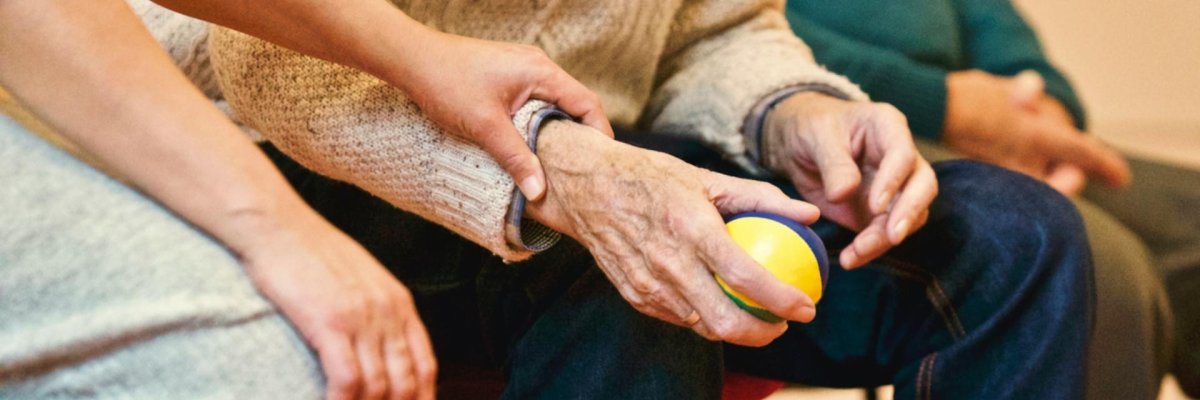 Foto. Hände eines alten Menschen, die einen kleinen Ball halten. Auf seinem Arm ruht die Hand einer jungen Person.