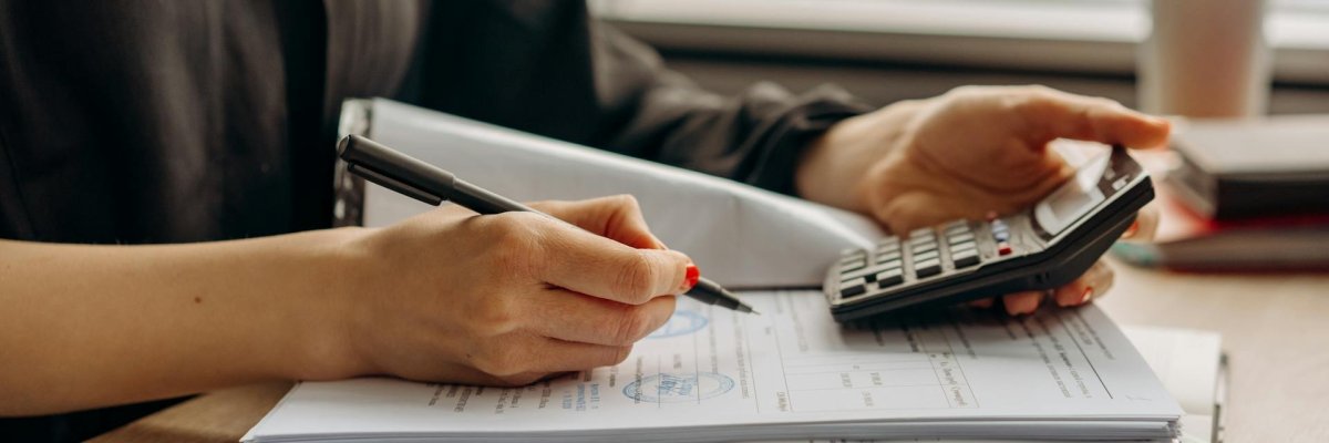 Foto: Aktenordner, Taschenrechner und eine Hand, die einen Stift hält.