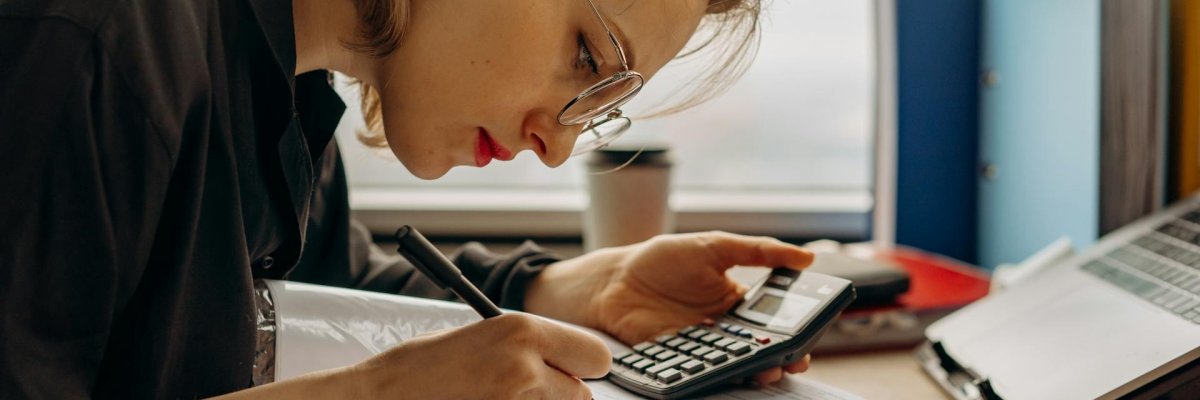 Foto: Eine über einen Aktenordner gebeugte Frau, die mit Hilfe eines Taschenrechners etwas berechnet.