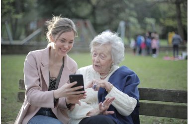 Foto: Eine junge Frau zeigt einer älteren Frau etwas auf ihrem Handy. Die beiden sitzen in einem Park auf einer Bank.