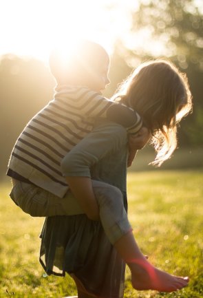 Ein älteres Kind trägt ein jüngeres Kind huckepack auf einer sonnigen Wiese. Das helle Gegenlicht der Sonne umrahmt die beiden und erzeugt eine warme, leichte Atmosphäre.