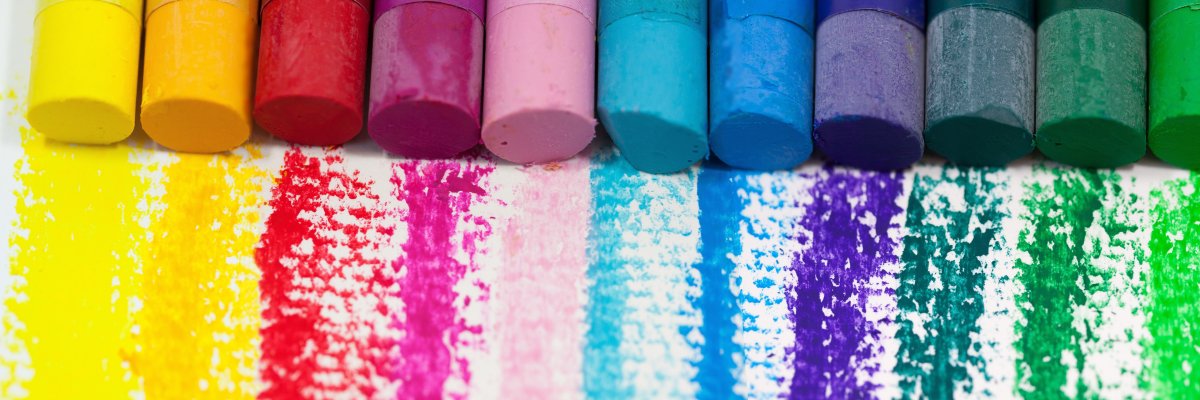 Eine Reihe farbiger Wachsmalstifte oder Pastellkreiden in einem breiten Farbspektrum, liegt nebeneinander. Darunter sind auf weißem Untergrund die kräftigen Farbstriche der jeweiligen Farben zu sehen. Dieses Bild symbolisiert die Vielfalt der Lebenswege und die bunten Entwicklungschancen, die durch Pflegefamilien und Adoption für Kinder geschaffen werden.