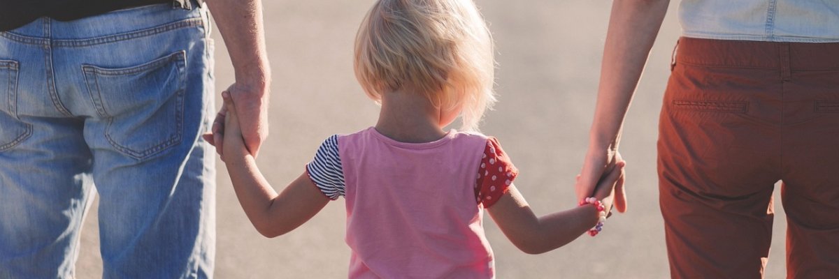 Ein kleines blondes Kind, von hinten gesehen, geht händchenhaltend zwischen zwei Erwachsenen. Das Bild symbolisiert die Fürsorge, Geborgenheit und den Halt, den Pflegeeltern einem Kind auf seinem Lebensweg geben.