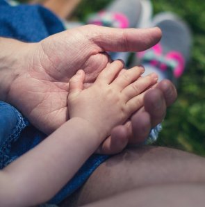 Eine Nahaufnahme zeigt eine kleinere Kinderhand, die sicher und geborgen in der Hand eines Erwachsenen liegt. Im unscharfen Hintergrund ist grüne Wiese zu erkennen.