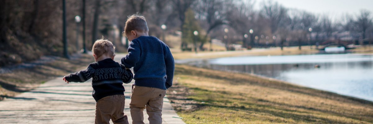 Zwei Kinder, von hinten gesehen, gehen gemeinsam auf einem Weg entlang eines sonnigen Seeufers. Das größere Kind scheint das kleinere zu begleiten oder zu führen. Dieses Bild symbolisiert die Unterstützung und Begleitung, die der Allgemeine Soziale Dienst des Jugendamts Familien und Kindern auf ihrem Lebensweg bietet.
