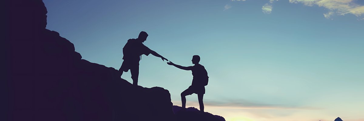 Silhouette zweier Personen auf einem felsigen Bergpfad. Die eine Person reicht der anderen unterstützend die Hand. Der Himmel ist in den Farbtönen des Sonnenaufgangs gehalten, was einen Neubeginn oder eine neue Perspektive symbolisiert.